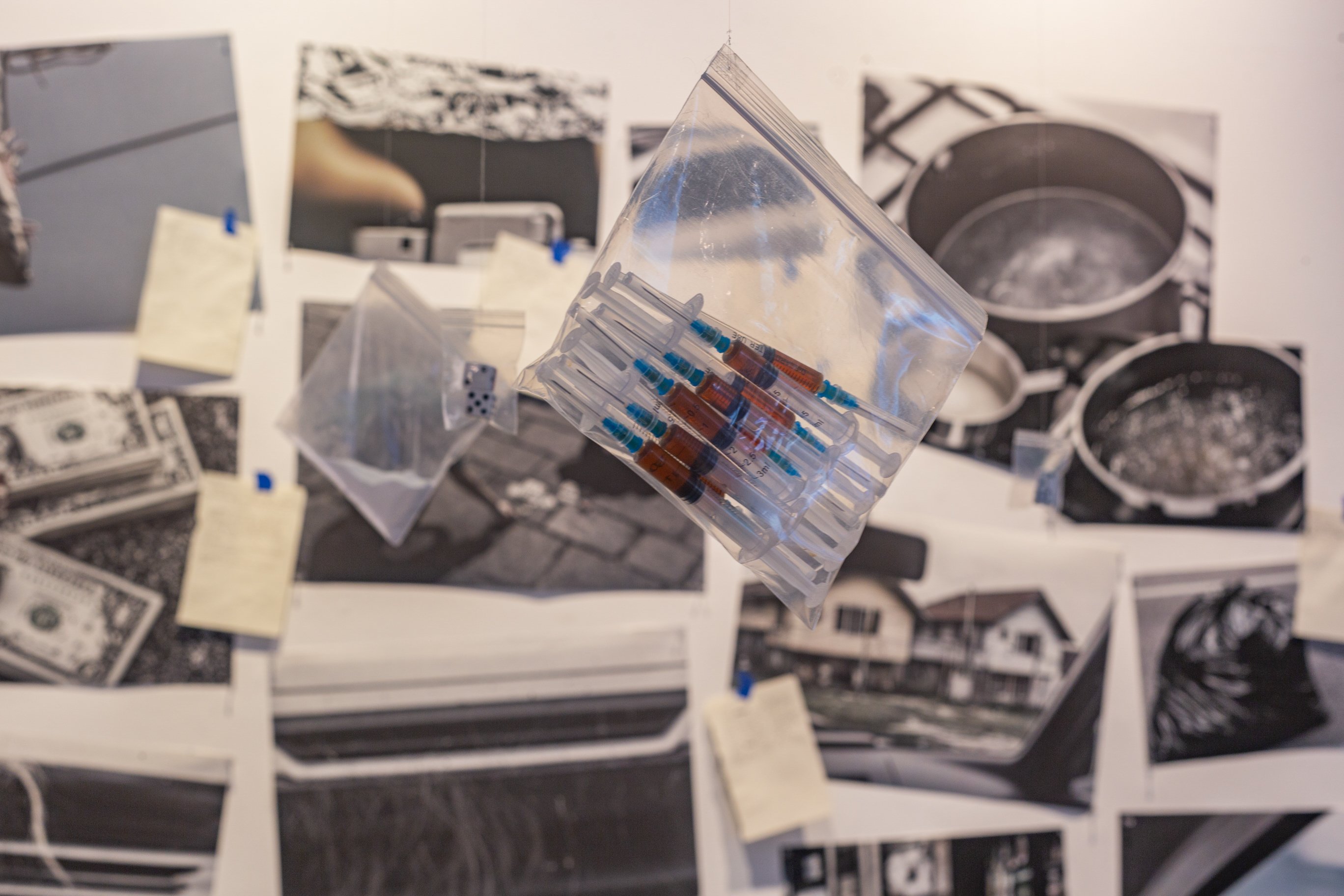 In the foreground, a ziplock bag with multiple needles, hangs suspended from the cieling. In the background are several photographic prints pasted on a white wall.